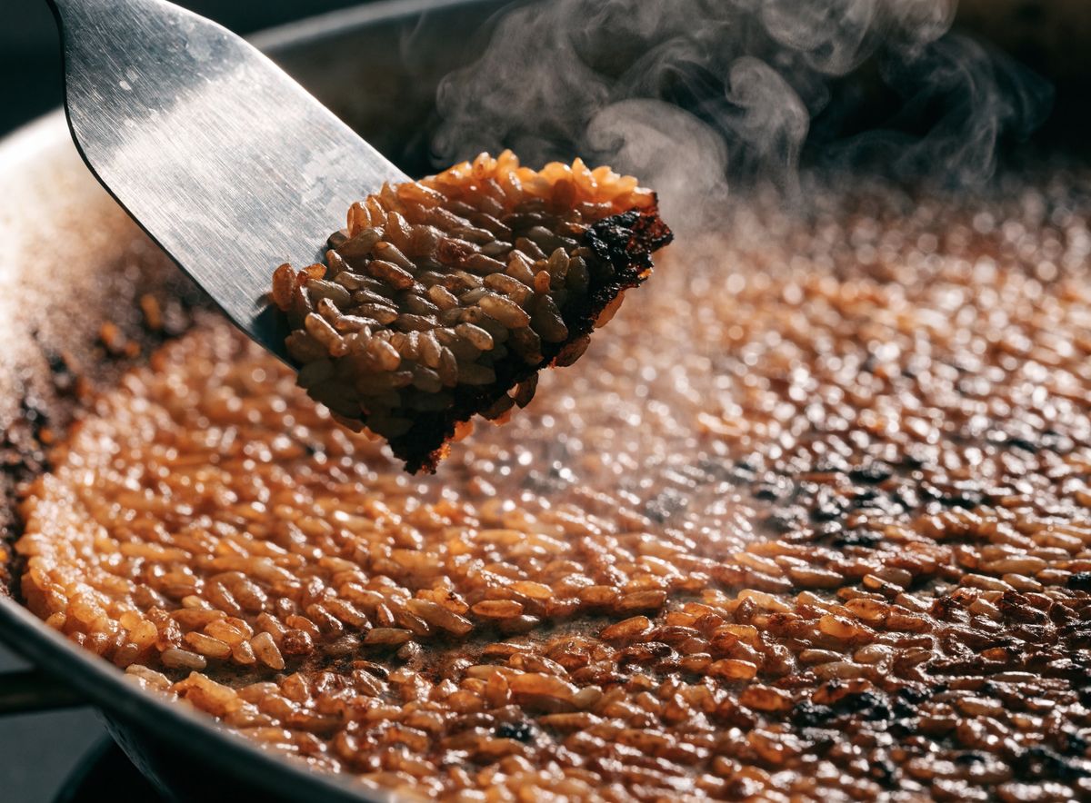 Cận cảnh thìa kim loại nạy lớp cơm cháy vàng nâu giòn ở đáy chảo paella